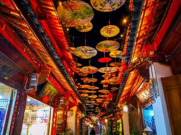 Night view of Lijiang Ancient Town, Yunnan, China – illuminated Naxi wooden houses, red lanterns, and cobbled streets reflecting in canals, with Jade Dragon Mountain silhouette. UNESCO-listed old town blending Dongba culture and romantic atmosphere.