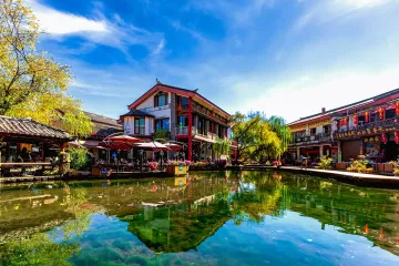 Scenic view of Shuhe Ancient Town in Lijiang, Yunnan, China - traditional Naxi architecture with stone bridges, flowing canals, and snow-capped Jade Dragon Mountain in background. UNESCO World Heritage Site showcasing well-preserved Tea Horse Road history.